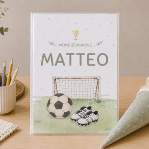 Könnte beinhalten: Ein personalisierter Ordner zum Thema Fußball mit dem Namen "MATTEO" in grünen Buchstaben. Das Cover zeigt einen Fußball, ein Tor und Stollen auf einem grünen Feld. Eine Trophäe und die Worte "MEINE ZEUGNISSE" stehen oben.