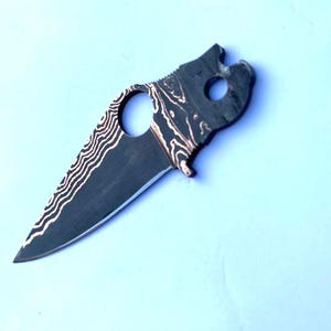 Può includere: Una lama di coltello personalizzata con un motivo unico. La lama ha una finitura scura e strutturata con un motivo ondulato a contrasto. Presenta un foro per le dita e una forma distintiva, probabilmente per un coltello pieghevole. La lama è su sfondo azzurro.