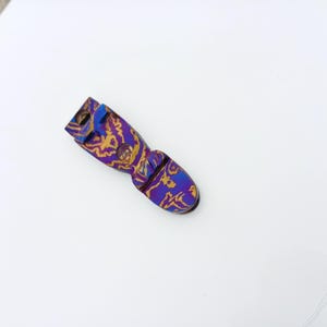 May include: A small, elongated metal component featuring a vibrant, iridescent finish with a swirling pattern of deep purple and metallic gold. The object has a rounded end and a structured, notched top section with two circular openings for attachment.