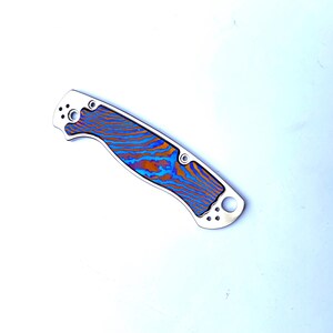 May include: A folding pocket knife with a stainless steel handle and a blade featuring a vibrant pattern of blue and orange swirls. The knife has a curved handle with multiple holes and a circular opening.
