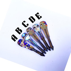 May include: Five metal tools with a colourful, patterned finish, arranged diagonally on a white surface. The tools have a pointed end and a rounded top with holes. The letters A, B, C, D, and E are printed above the tools.