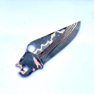 Op de afbeelding: Een op maat gemaakt mes met een donker, getextureerd lemmet met koperen accenten en een uniek patroon. Het mes heeft een gat bij het handvat en een kenmerkende vorm, ideaal voor verzamelaars of outdoorliefhebbers.