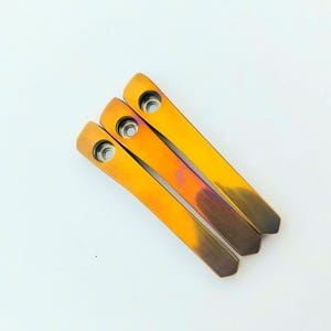 Puede incluir: Tres objetos metálicos alargados con un degradado de colores que va del oro al bronce. Cada uno tiene un agujero circular en un extremo. Los objetos están dispuestos en diagonal sobre un fondo blanco.