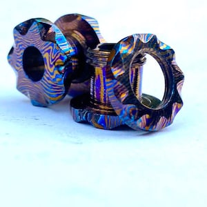 May include: Close-up of three pieces of jewelry with a unique, patterned design. The pieces are primarily blue, purple, and gold, with a textured, layered appearance. The jewelry has a gear-like shape, with a central ring and two circular pieces.