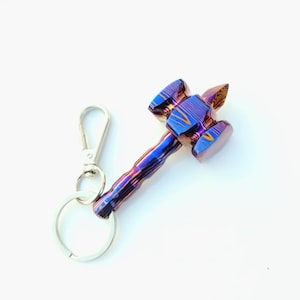 May include: A keychain featuring a hammer-shaped pendant with a multi-colored, iridescent, layered metal finish in shades of blue, purple, and copper. The pendant is attached to a standard silver-colored metal split ring and a lobster claw clasp.