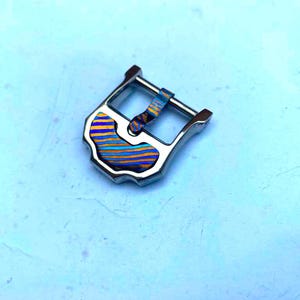 Puede incluir: Hebilla de reloj plateada con incrustación decorativa. La incrustación presenta un patrón de rayas azules, naranjas y amarillas. La hebilla tiene forma rectangular con un diseño curvo y una barra para la correa del reloj.
