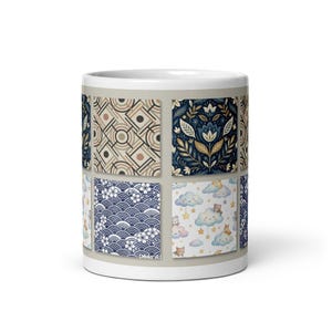 May include: A white ceramic mug with a collage of patterned designs. The patterns include floral, geometric, and wave motifs in blue, beige, and gold. The mug has a standard handle and a glossy finish.