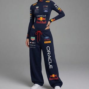 Puede incluir: Conjunto de body y pantalón de chándal azul marino con los logotipos de Red Bull y Oracle. El body de manga larga tiene cuello alto. El pantalón de chándal tiene cintura con cordón y la palabra "ORACLE" impresa verticalmente en la pierna.