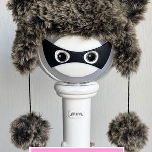 May include: A plush, faux fur hat in shades of gray, with ear flaps and pom-poms. The hat is placed on a white, cylindrical object with a black mask and large eyes. Text on a pink background reads "PDF Crochet Pattern."