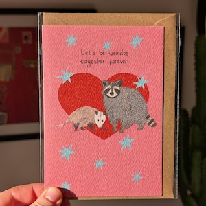 May include: A pink greeting card with a red heart and the text "Let's be weirdos together forever." The card features illustrations of a raccoon and an opossum. Blue star accents surround the heart. The card is in a clear plastic sleeve.