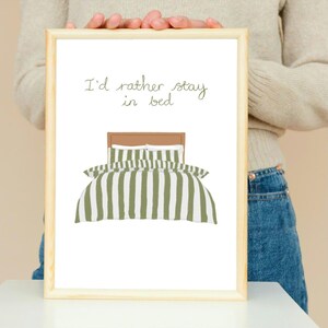 May include: A framed print featuring the phrase "I'd rather stay in bed." The artwork shows a bed with a brown headboard and green and white striped bedding. The frame is made of light-coloured wood.