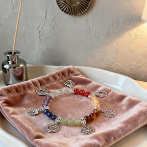 Op de afbeelding: Een chakra-armband met kleurrijke edelsteen kralen en zilveren bedels. De armband heeft een spectrum aan kleuren, waaronder rode, oranje, gele, groene, blauwe, paarse en heldere kralen. De bedels hebben ingewikkelde ontwerpen. De armband wordt getoond op een roze fluwelen zakje.