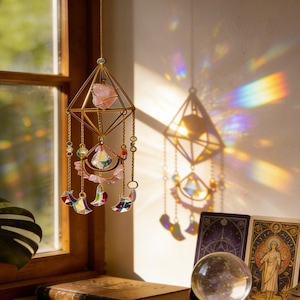 May include: A hanging gold-colored geometric prism suncatcher with a pink crystal and dangling iridescent crystals. The suncatcher is suspended in front of a window, casting rainbow light patterns on the wall. Tarot cards and a crystal ball are visible.