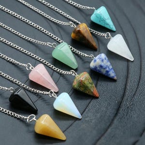 Könnte beinhalten: Eine Sammlung von Edelstein-Pendeln mit Silberketten. Die Pendel sind in verschiedenen Farben erhältlich, darunter Türkis, Grün, Rosa und Schwarz. Jedes Pendel hat eine spitze, konische Form. Die Ketten sind verbunden.