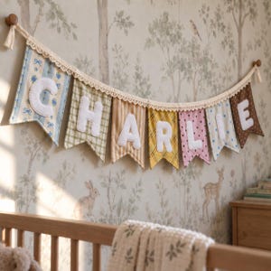 May include: A decorative fabric banner featuring seven pennants hanging from a cream-coloured lace trim. Each pennant displays a large white felt letter spelling the name CHARLIE, with each flag showcasing a unique pattern in blue, green, tan, yellow, pink, and brown.