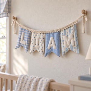 Puede incluir: Un banderín de tela con cuatro gallardetes en tonos azul y blanco, que deletrean el nombre LIAM con letras de tela color crema. Los gallardetes muestran patrones de cuadros vichy, estrellas, rayas y flores, con un borde de encaje crema.