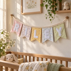 Puede incluir: Una guirnalda de tela con cinco banderines en colores pastel, cada uno con una letra blanca para formar el nombre RILEY. Los banderines presentan patrones de color rosa, floral, cuadros amarillos, rayas azules y lunares verdes con encaje.