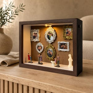 May include: A rectangular dark wood shadow box display case featuring an internal light, five ornate picture frames in gold and silver tones containing couple photographs, and several small human figurines standing on the white base surface.