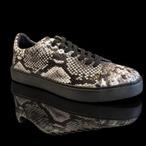 Peut inclure: Une basket basse avec un motif peau de serpent noir et blanc sur la tige, des lacets noirs et une semelle en caoutchouc noir uni. La chaussure est présentée sur un fond noir avec un reflet net visible sous la base de la basket.
