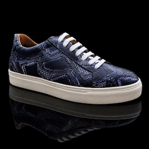 Könnte beinhalten: Ein Low-Top-Sneaker mit dunkelblauem Obermaterial und strukturiertem Schlangenledermuster. Der Schuh verfügt über weiße Schnürsenkel, ein beiges Innenfutter und eine dicke, strukturierte weiße Gummisohle mit einer abgerundeten Schuhspitze.
