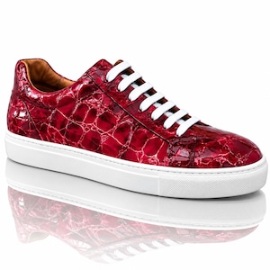 Könnte beinhalten: Ein Low-Top-Sneaker mit einem glänzenden, tiefroten Obermaterial in einer strukturierten, rissigen Lederoptik. Der Schuh verfügt über strahlend weiße Schnürsenkel und eine dicke, durchgehende weiße Gummisohle. Das Innenfutter ist aus beigem Material.