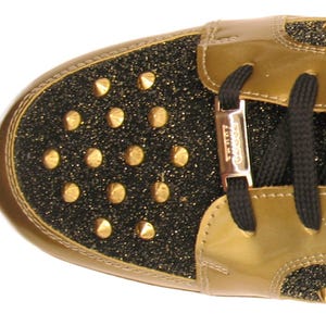 Puede incluir: Primer plano de una zapatilla dorada y negra. La puntera y el tal&oacute;n son negros con tachuelas doradas. La parte superior es de charol dorado. Los cordones negros est&aacute;n ensartados en una ojal met&aacute;lico dorado.