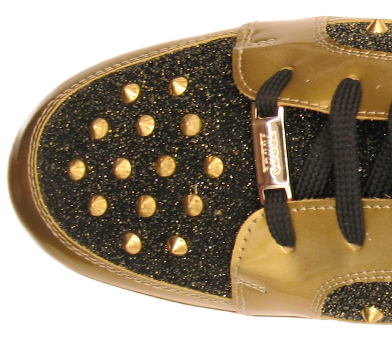 Puede incluir: Primer plano de una zapatilla dorada y negra. La puntera y el tal&oacute;n son negros con tachuelas doradas. La parte superior es de charol dorado. Los cordones negros est&aacute;n ensartados en una ojal met&aacute;lico dorado.