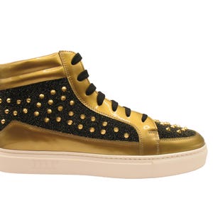 Puede incluir: Zapatilla alta dorada y negra. La zapatilla presenta una parte superior de charol dorado con detalles de purpurina negra y tachuelas doradas. Cordones negros y suela blanquecina.