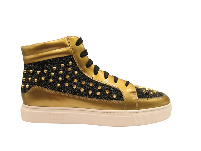 Puede incluir: Zapatilla alta dorada y negra. La zapatilla presenta una parte superior de charol dorado con detalles de purpurina negra y tachuelas doradas. Cordones negros y suela blanquecina.