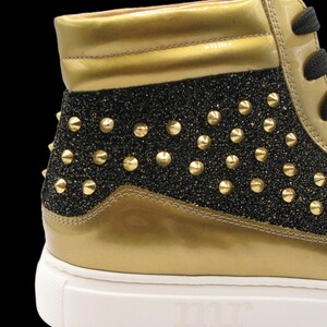 Puede incluir: Zapatilla alta con detalles dorados y negros con purpurina. El zapato presenta tachuelas doradas y una suela blanca con las letras "mr" en relieve. La parte superior del zapato es dorada, con un panel negro con purpurina.