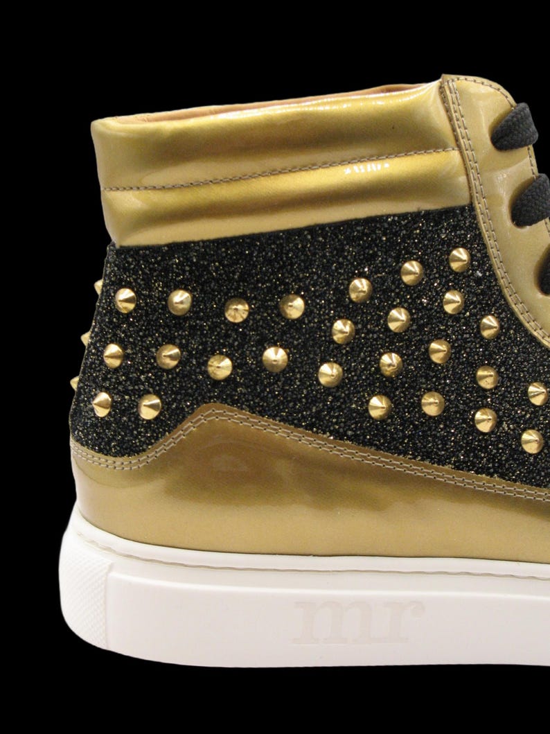 Puede incluir: Zapatilla alta con detalles dorados y negros con purpurina. El zapato presenta tachuelas doradas y una suela blanca con las letras "mr" en relieve. La parte superior del zapato es dorada, con un panel negro con purpurina.