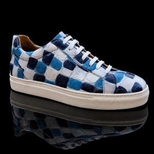 Könnte beinhalten: Ein Low-Top-Sneaker mit einem glänzenden, strukturierten Obermaterial in einem Karomuster aus Weiß, Hellblau und Dunkelmarineblau. Der Schuh verfügt über weiße Schnürsenkel, ein beiges Innenfutter und eine durchgehende weiße Gummisohle.