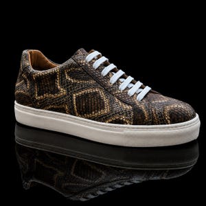 Könnte beinhalten: Ein Low-Top-Sneaker mit einem braun-schwarzen Schlangenledermuster und strukturierter Oberfläche. Der Schuh verfügt über weiße Schnürsenkel und eine dicke, durchgehend weiße Gummisohle. Das Innenfutter ist in einem hellen Beigeton gehalten.
