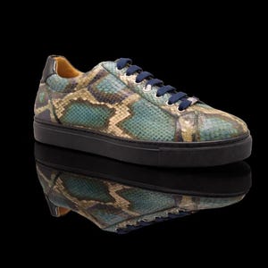 Könnte beinhalten: Ein Low-Top-Sneaker mit einem Obermaterial in Schlangenlederoptik in Blaugrün und Beige, dunkelblauen Schnürsenkeln, silberfarbenen Ösen und einer durchgehend schwarzen Gummisohle. Das Innenfutter ist hellbraun und die Fersenkappe dunkelblau.