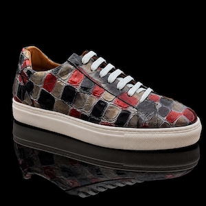 Könnte beinhalten: Ein Low-Top-Sneaker mit einem glänzenden, strukturierten Obermaterial mit einem sich wiederholenden Muster aus schwarzen, roten und metallisch silbernen rechteckigen Feldern. Der Schuh hat weiße Schnürsenkel, eine weiße Gummizwischensohle und ein beiges Innenfutter.