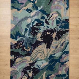 May include: A rectangular area rug with an abstract design in shades of teal, green, purple, and beige. The rug's pattern resembles a landscape or topographical map, with swirling shapes and color variations. The rug is placed on a light-colored wooden floor.