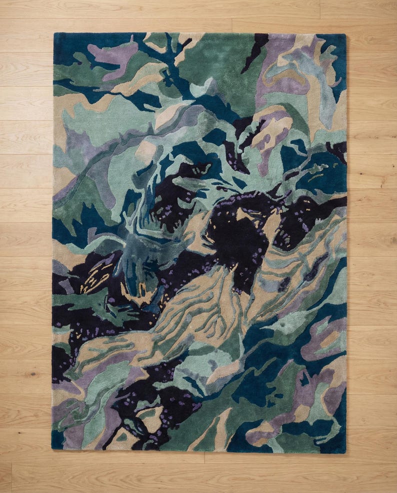 May include: A rectangular area rug with an abstract design in shades of teal, green, purple, and beige. The rug's pattern resembles a landscape or topographical map, with swirling shapes and color variations. The rug is placed on a light-colored wooden floor.