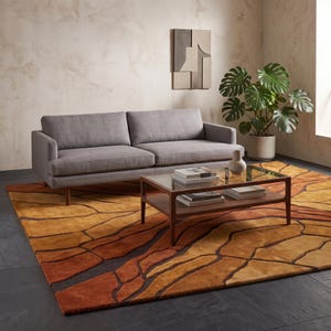 May include: A gray sofa with two cushions and wooden legs sits on a large area rug featuring an abstract pattern of orange, brown, and yellow shapes. A rectangular coffee table with a glass top and wooden frame holds books and a small vase.