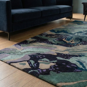 May include: A large area rug with an abstract design in shades of blue, green, purple, and beige. The rug is placed on a hardwood floor, with a navy blue sofa in the background. The rug's pattern resembles a landscape.