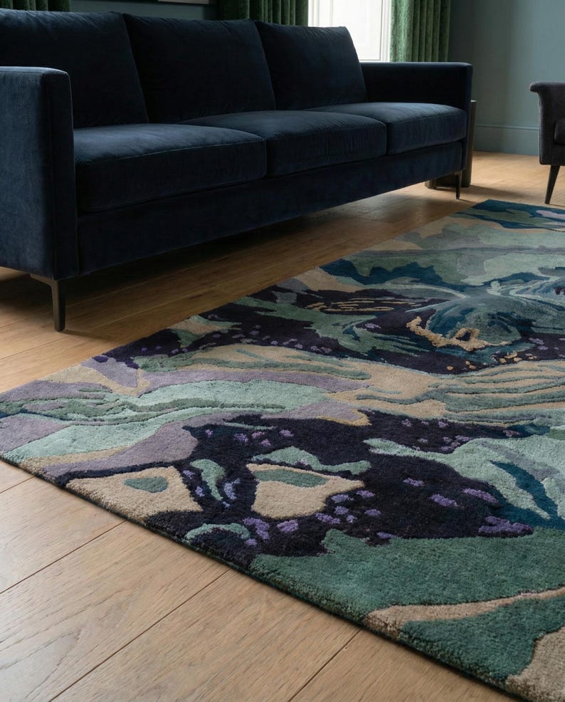 May include: A large area rug with an abstract design in shades of blue, green, purple, and beige. The rug is placed on a hardwood floor, with a navy blue sofa in the background. The rug's pattern resembles a landscape.