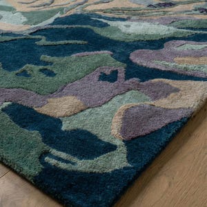 May include: A close-up of a patterned area rug with a landscape design. The rug features shades of teal, green, beige, and purple. The rug is made of a soft, plush material and is placed on a wooden floor.