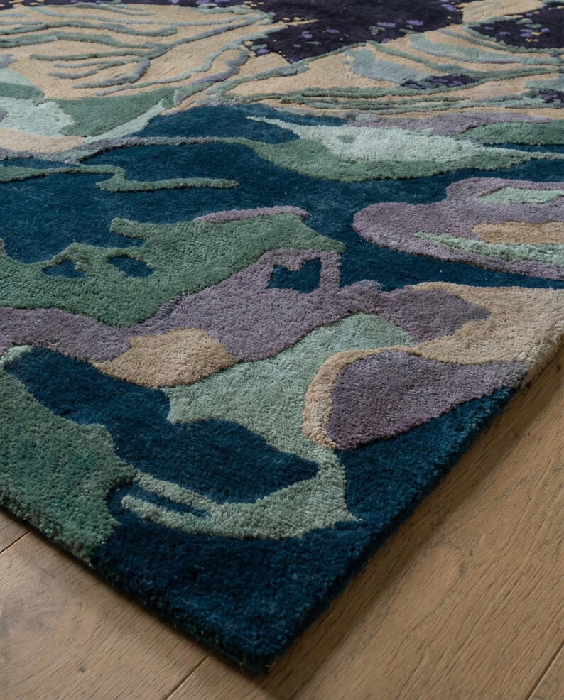 May include: A close-up of a patterned area rug with a landscape design. The rug features shades of teal, green, beige, and purple. The rug is made of a soft, plush material and is placed on a wooden floor.