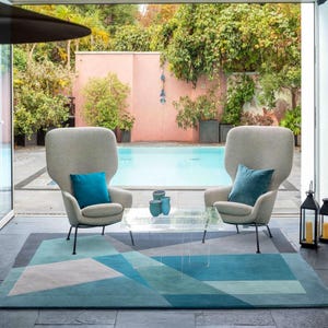 Op de afbeelding: Twee hooggerugde, lichtgrijze fauteuils met teal kussens staan tegenover elkaar aan een heldere glazen tafel. Een geometrisch tapijt in blauw- en grijstinten ligt eronder. De sc&egrave;ne speelt zich af in een buitenruimte met een zwembad.