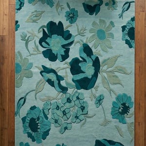K&ouml;nnte beinhalten: Ein rechteckiger Teppich mit floralem Muster. Der Teppich zeigt gro&szlig;e, stilisierte Blumen in T&uuml;rkis- und Dunkelt&uuml;rkist&ouml;nen auf hellblauem Hintergrund. Der Teppich liegt auf einem Holzboden.