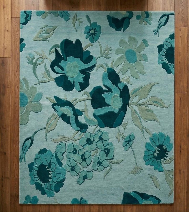 K&ouml;nnte beinhalten: Ein rechteckiger Teppich mit floralem Muster. Der Teppich zeigt gro&szlig;e, stilisierte Blumen in T&uuml;rkis- und Dunkelt&uuml;rkist&ouml;nen auf hellblauem Hintergrund. Der Teppich liegt auf einem Holzboden.