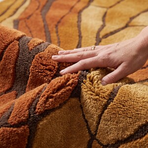 May include: A thick, tufted rug featuring a textured, abstract pattern in shades of burnt orange, mustard yellow, and deep brown. The surface has a high-pile, plush construction with distinct carved lines separating the colored sections.