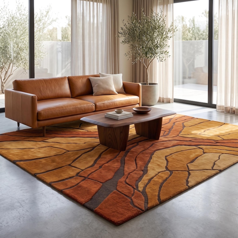 May include: A large rectangular area rug featuring a textured, abstract pattern of organic, cracked earth shapes in shades of burnt orange, mustard yellow, and deep brown, laid out on a smooth concrete floor beneath a leather sofa and coffee table.