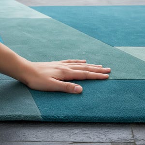 Op de afbeelding: Een close-up van een geometrisch patroon tapijt in blauw en turquoise. Het tapijt heeft een zacht, getextureerd oppervlak met verschillende tinten blauw en turquoise. Een hand rust op het tapijt, wat de textuur en kleurvariaties benadrukt.