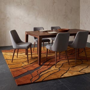 May include: A rectangular dark wood dining table with four gray upholstered chairs featuring tapered wooden legs. The table is set with white plates and clear glasses. The furniture sits on a large area rug with a textured, abstract pattern in shades of orange, yellow, and brown, placed on a dark gray stone floor.