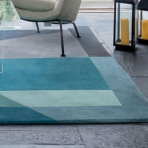 Op de afbeelding: Een rechthoekig vloerkleed met een geometrisch patroon in blauw- en grijstinten. Het kleed heeft een centraal turquoise gedeelte, omlijst door lichtere blauwe en grijze panelen. Het ligt op een stenen vloer, met een stoel en een lantaarn op de achtergrond.